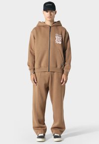 Brauner Reißverschluss-Hoodie mit Kängurutaschen und grafischem Druck. Passende braune Hose mit weißer Naht und schwarzen Schuhen.
