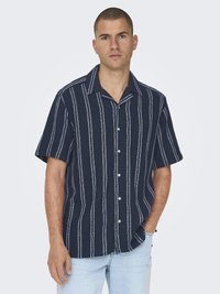 Chemise bleu marine à manches courtes avec des rayures blanches verticales, boutons sur le devant, tissu texturé et coupe décontractée. Associée à un jean bleu clair.