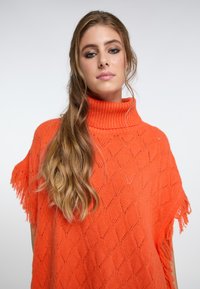 Poncho de punto naranja con cuello alto acanalado, que presenta patrones de diamante abiertos y bordes con flecos. Textura suave, corte relajado.