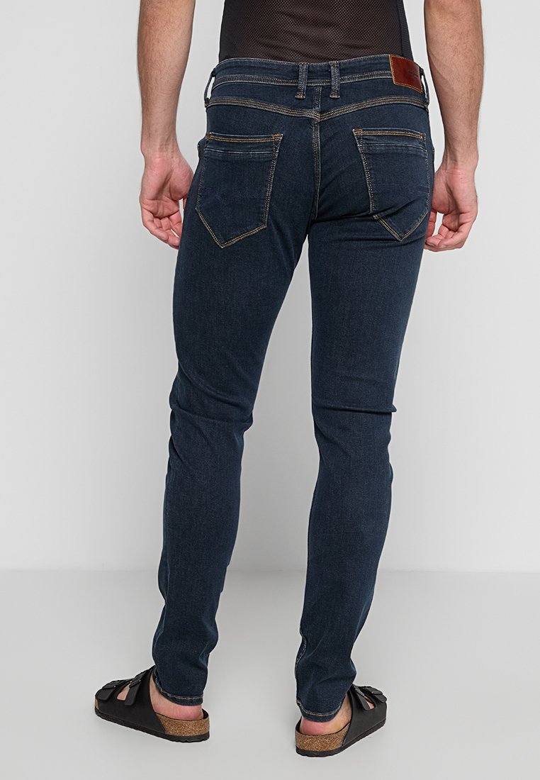 Eine Person, die dunkelblaue enge Jeans und schwarze Sandalen trägt, wird von hinten gezeigt, mit den Händen an den Seiten, vor einem schlichten Hintergrund.