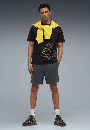 Junger Mann trägt schwarzes T-Shirt mit gelber Cartoon-Grafik, graue Shorts, schwarze Sneakers, weiße Socken und einen gelben Pullover lässig über die Schultern gelegt.