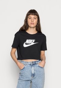 Camiseta negra cortada de algodón con un logo de Nike blanco. Presenta mangas cortas y un cuello redondo. Combinada con unos jeans de mezclilla azul claro.