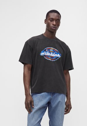 Άνδρας που φοράει μαύρο t-shirt Dickies και ανοιχτόχρωμο τζιν, στέκεται μπροστά σε μια μονόχρωμη ανοιχτογκρι φόντο.