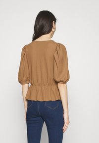 Blusa marrón con textura que presenta mangas abullonadas y un dobladillo con volantes, ajustada en la cintura para un look entallado. Combinada con jeans oscuros.