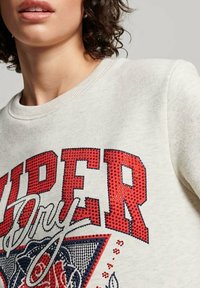 Kobieta ubrana w jasnoszary sweter z dużymi, czerwonymi, ćwiekowanymi literami oraz dekoracyjnym napisem na klatce piersiowej.