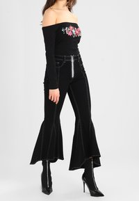 Schwarzes Off-Shoulder-Top mit floraler Stickerei, kombiniert mit schwarzen Schlagjeans mit Kontrastnähten und metallischen Reißverschlüssen.