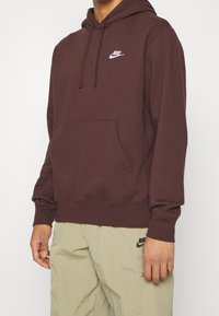 Mörkröd Nike-hoodie med framficka, justerbar dragsko på huvan och vit logotyp på bröstet. Mjukt tyg, avslappnad passform med ribbade mudd.