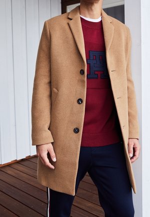 Mand iført en kamelfarvet uldfrakke over en rød sweater med et stort mørkeblåt "H"-logo og mørkeblå bukser med hvide og røde striber.