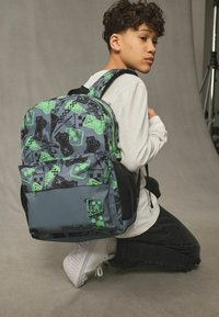 Mochila con un tejido gris y diseños verdes de Minecraft, que incluye personajes y patrones. Bolsillos laterales de malla negra y correas ajustables.
