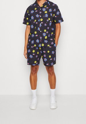 Korte mouw zwarte bloemen blouse en bijpassende shorts met gele glimlachgezichten, blauwe bloemen en rode accenten. Witte sneakers maken de look compleet.