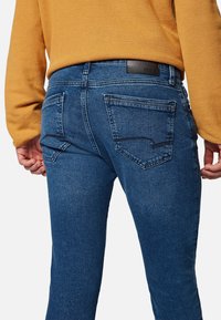 Los jeans de mezclilla azules cuentan con un diseño ajustado, cinco bolsillos y un parche de cuero negro en la parte trasera de la cintura. La tela tiene una textura suave.
