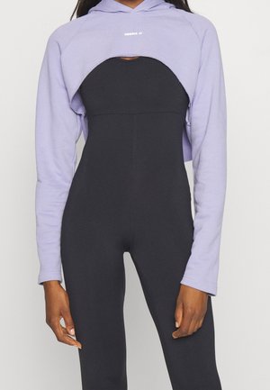 Frau trägt einen kurzen lavendelfarbenen Hoodie über einem enganliegenden schwarzen ärmellosen Overall und steht vor einem schlichten hellen Hintergrund.