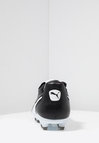 Bota de fútbol en blanco y negro con un upper de cuero texturizado, forma curva, suela moldeada y parche de logo prominente en el talón.