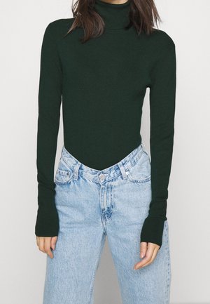 Femme portant un pull à col roulé vert foncé et un jean taille haute bleu clair à coupe décontractée, debout devant un fond uni.