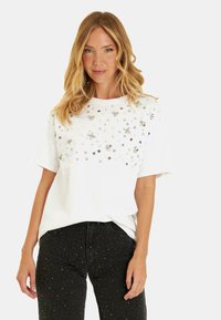 T-shirt blanc à col rond, orné de sequins argentés et de motifs floraux sur le devant. Associé à un jean noir avec des détails à clous.