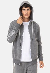Graue Zip-Hoodie mit Kapuze zum Zugbinden, mit einem weißen Grafikdesign auf dem linken Ärmel und kontrastierenden wei�ßen Reißverschlussdetails.