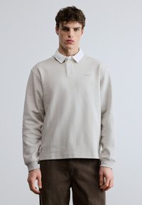 Tricou polo cu mâneci lungi de culoare gri deschis, cu guler alb, având un logo subtil pe piept și manșete cu nervuri, asortat cu pantaloni maro închis.