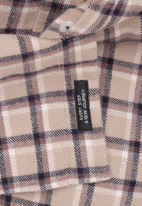 Beige en bruine geruite flanellen stof met witte en marineblauwe accenten. Heeft een zwart etiket met de tekst: "IEDERE STAP IS EEN NIEUWE REIS."