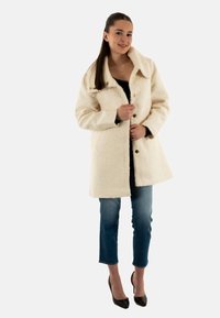 Abrigo de lana texturizada blanca con cuello, botones en la parte frontal y dos bolsillos, combinado con jeans de mezclilla azules y tacones altos negros.