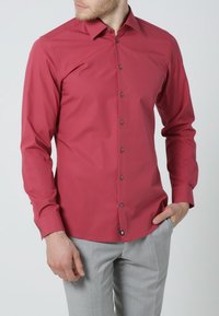 Rotes Langarmhemd mit figurbetontem Schnitt, Knopfleiste am Kragen und grauen Knöpfen, kombiniert mit hellgrauer Hose. Glatter, leicht glänzender Stoff.