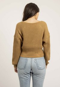 Pull en tricot de couleur marron chaud avec une coupe ample, un col large, un ourlet côtelé et des manches longues, associé à un jean bleu clair.