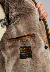 Bruine nepbont binnenkant van een jas met een oranje voering en een label waarop "SNOWPACK ISSUE 409/33" staat. Rits- en knoopdetails zijn zichtbaar.