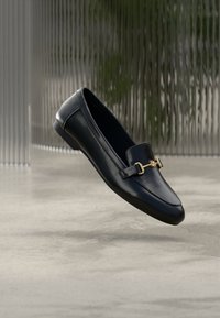 Mocassin en cuir noir à finition lisse, orné d'un détail en métal doré et d'un bout arrondi, suspendu sur un fond minimaliste.