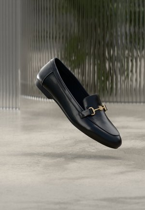 Svart lærloafer med glatt finish, med gullfarget maskinvareaccent og avrundet tå, hengende mot en minimalistisk bakgrunn.