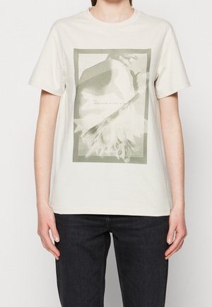 Osoba ubrana w kremowy T-shirt z krótkim rękawem z subtelnym kwiatowym nadrukiem oraz tekstem "Nineteen Sixty Eight" i "Calvin Klein" na przedzie.