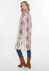 Langer weißer Cardigan mit roten geometrischen Mustern, kombiniert mit dunkelblauen Jeans und braunen High Heels. Glatte Textur, lockere Passform.