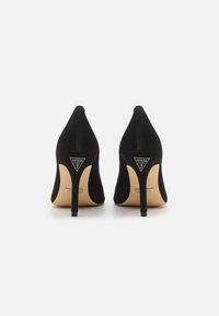 Scarpe col tacco alto in suede nero con punta affilata, caratterizzate da un sottile tacco a stiletto e un logo argentato sul retro. Texture liscia e design elegante.