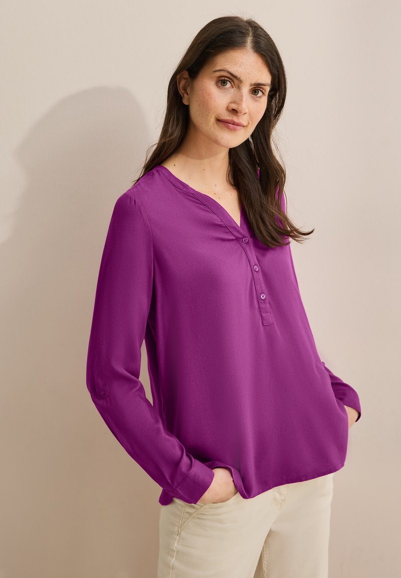 Blusa de manga larga morada con escote en V, con botonera y textura suave, llevada sobre pantalones de color claro.