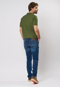 Polo vert à manches courtes, associé à un jean bleu. Les chaussures du modèle présentent un dessus marron et une semelle crème. La vue de dos montre un surpiqûre détaillée.
