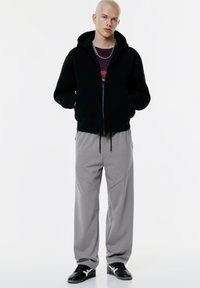 Sort zip-up hættejakke med hætte, parret med løse grå bukser med paneldetaljer, båret med sorte sneakers. Simpel, atletisk stil.
