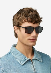 Occhiali da sole quadrati neri con lenti scure, caratterizzati da dettagli in metallo e un design elegante. Indossati da una persona con una giacca di jeans azzurro chiaro.