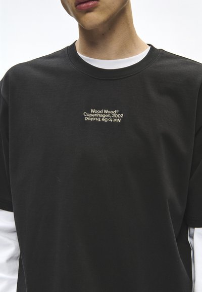 Νέος/νεαρή φοράει μαύρο t-shirt Wood Wood πάνω από λευκό μακρυμάνικο πουκάμισο, με μικρό κείμενο "Wood Wood Copenhagen, 2002 Not to be trusted" στο στήθος.