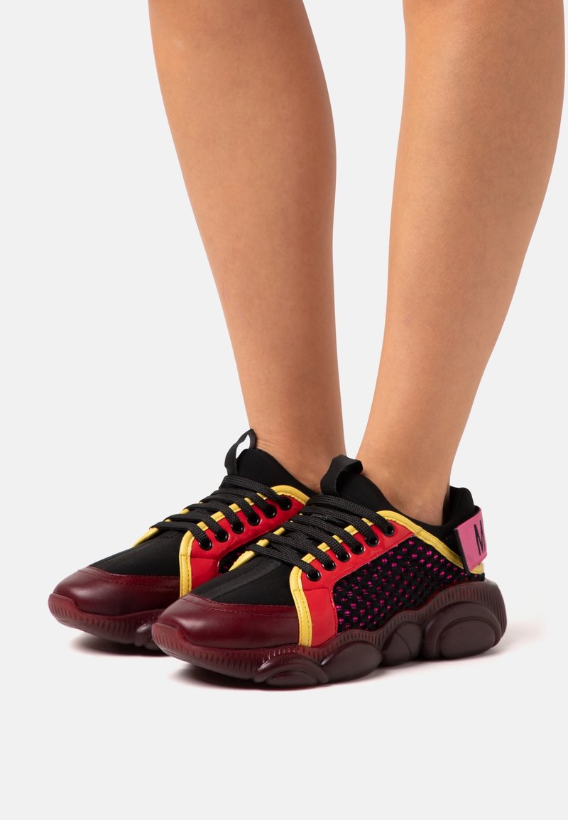 Jambes portant des baskets noires avec des détails rouges, jaunes et roses et des semelles épaisses marron foncé sur fond blanc.