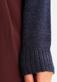 Marinblå stickad tröja med ärm och ribbad mudd, kombinerad med ett vinrött tyg; har en mjuk, texturerad yta.