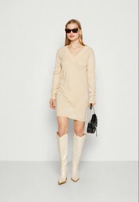 Kreminis džemperio stiliaus suknelė su V formos iškirpte ir ilgomis rankovėmis, derinama su baltomis iki kelių aukštakulnėmis botais ir maža juoda rankine. Lygus audinys, kasdieniškas dizainas.