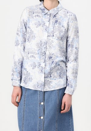 Biała koszula z długim rękawem z niebieskim botanicznym i krajobrazowym nadrukiem, zestawiona z niebieską spódnicą dżinsową zapinaną na guziki od frontu, noszona przez stojącą osobę.