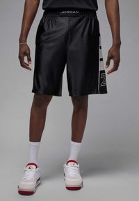 Shorts de basket-ball noirs avec une texture brillante, présentant un panneau latéral blanc avec des numéros et un logo à la taille. Portés avec des chaussettes et des chaussures blanches.