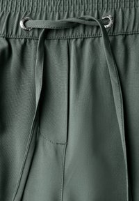 Tissu vert foncé, avec une taille élastique dotée d'un cordon de serrage, des accents en métal argenté, et une poche latérale. Texture lisse.