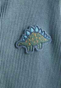 Vyšívanie zeleného stegosaura na textúrovanej svetlomodrej látke, s modrými tŕňmi a oranžovými bodkami na chrbte.