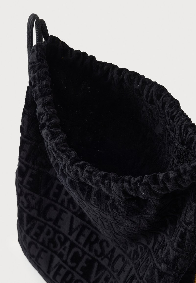 Sort bøtte-taske i fløjl med samlet top, prydet med præget Versace-logo mønster. Blød tekstur, rund form og snørelukning.