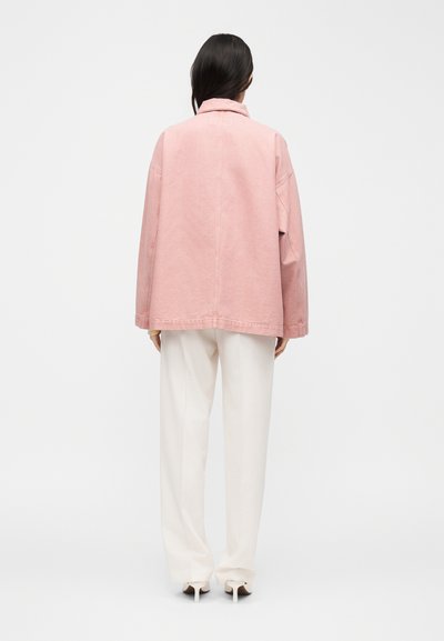 Γυναίκα με σκούρα μαλλιά που φοράει ένα φαρδύ ροζ σακάκι, λευκό παντελόνι καμπάνα και λευκές ψηλοτάκουνες γόβες, στέκεται με την πλάτη προς τον θεατή σε ένα απλό φόντο.