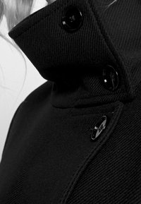 Sort uldfrakke med høj krave, to store sorte knapper og tekstureret stof med diagonale linjer.