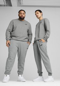 Grå sweatshirt og joggers lavet af blødt stof med ribbede manchetter. Har sorte logo accenter på brystet og låret. Hvide sneakers.