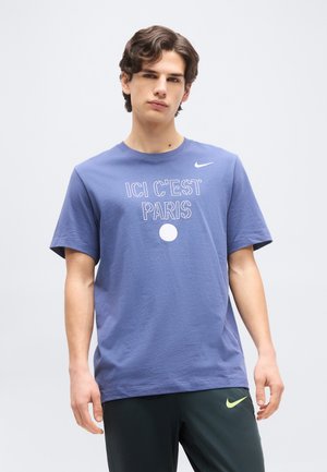 Jeune homme portant un t-shirt bleu Nike "ICI C'EST PARIS" et un pantalon Nike foncé, debout devant un fond clair uni.
