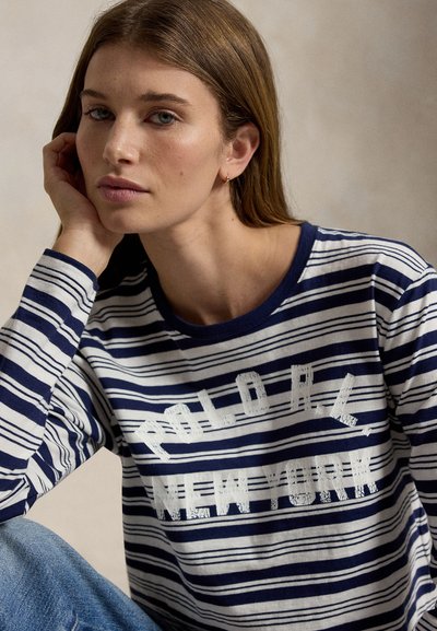 Femme aux cheveux châtain clair portant un t-shirt à manches longues rayé bleu marine et blanc avec le texte « Polo L.L. New York », reposant sa tête sur sa main.