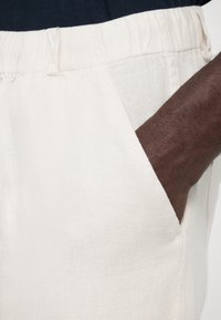 Světlé lněné šortky mají uvolněný střih s elastickým pasem. Vedle kapsy je umístěna tmavá ruka, která zdůrazňuje strukturu látky.